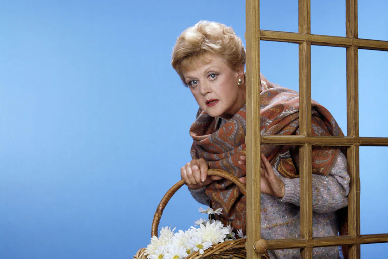 A woman holding a basket of flowers in a large scarf hides behind a door
