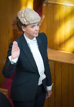 Woman in white wig and black gown holds up hand to swear oath.