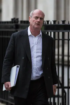 Man in black suit walking along a street with a white file under his arm.