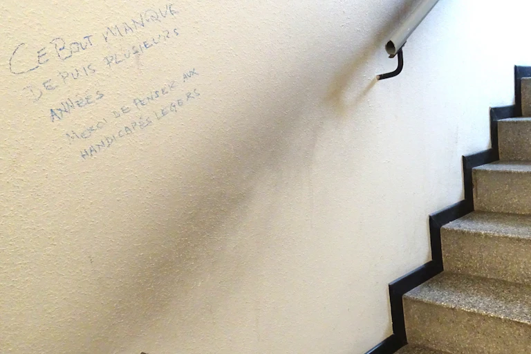 Graffiti dans un escalier à Genève, où il manque une section de main-courante : « Ce bout manque depuis plusieurs années / Merci de penser aux handicapés légers »