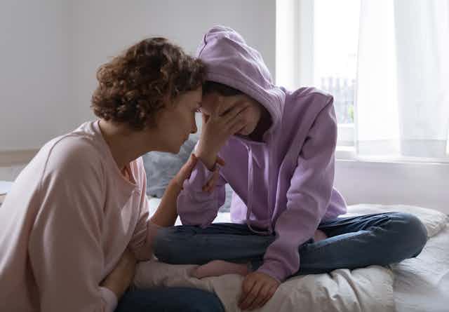Worried mom leaning forward to comfort her teen daughter who is sitting on her bed with her hand over her face.