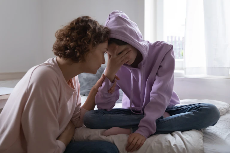 Worried mom leaning forward to comfort her teen daughter who is sitting on her bed with her hand over her face.