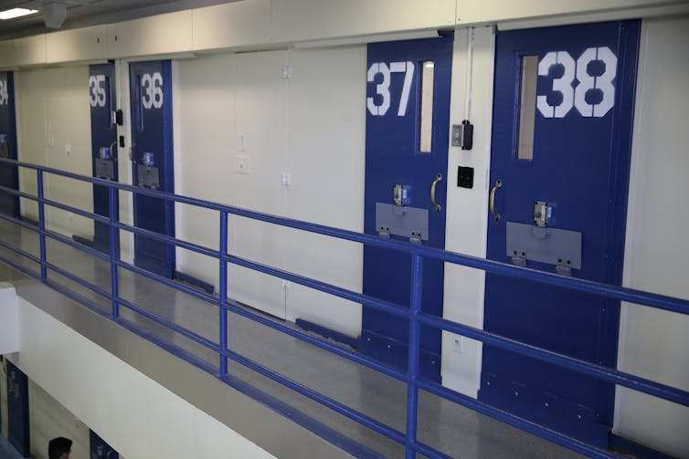 Blue pained doors with numbers in white paint on them are seen along a barren prison corridor.