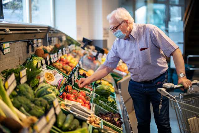 A white elderly man holds a cart in his left hand and with his right reaches out for tomatoes in a vegetable and fruit section