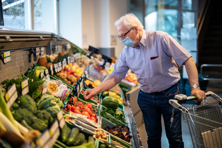 A white elderly man holds a cart in his left hand and with his right reaches out for tomatoes in a vegetable and fruit section