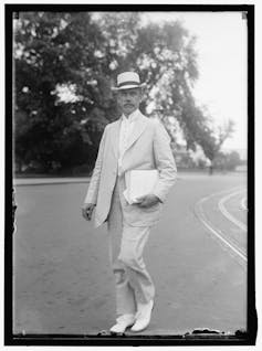A black and white photo shows a formally dressed man in a light-colored suit walking across a lawn.