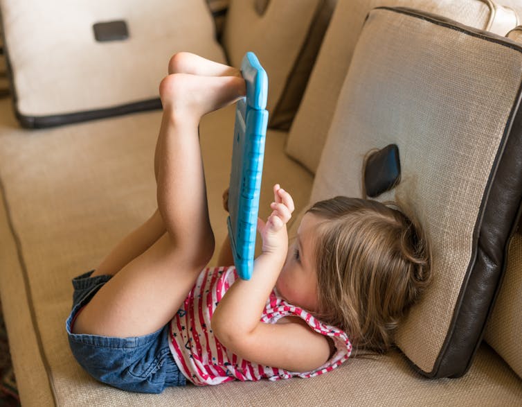 A small child holding an ipad up with their feet while on the sofa