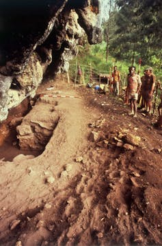 Some men stand to the side at the Nombe rockshelter, with parts of the earth on the bottom-left partially excavated.