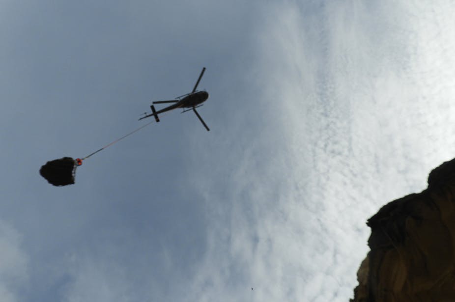 A helicopter is seen from below, silhouetted against a blue sky. A cable connects it to a large rock