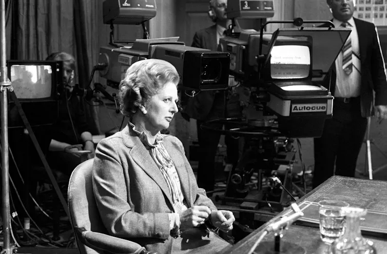 Margaret Thatcher sat at a desk with TV cameras behind her.
