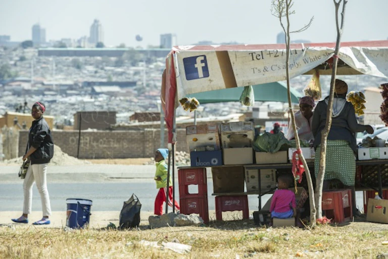 Roadside stall with crowded settlement in the background. Further away are tall buildings.