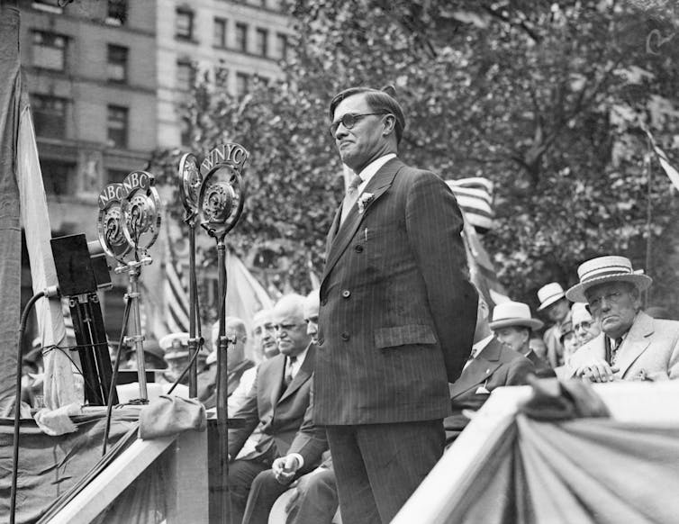 A white man dressed in a business suit is standing on stage as dozens of other white men sit in chairs and listen to his speech..