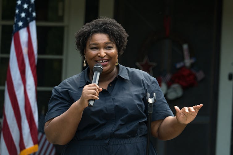 standing near an American flag, a black womanis giving a speech while holding a microphone with one hand and gesturing to the crowd with her other.