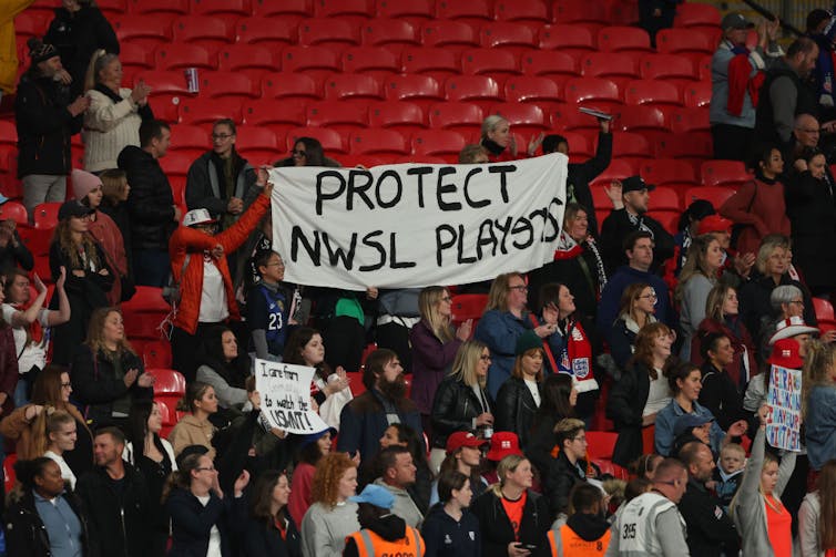 People in a stadium with red seats hold a banner reading 'Protect NWSL Players.'