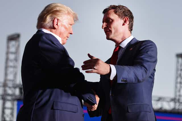 Two men in blue blazers, white shirts and red ties shaking hands.
