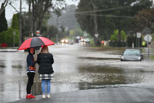 Two women under an umbrella look at a stranded car in a flooded road