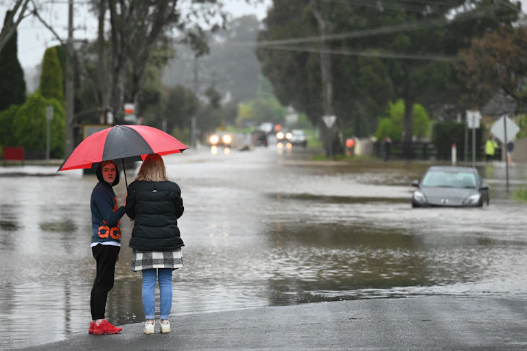 Two women under an umbrella look at a stranded car in a flooded road