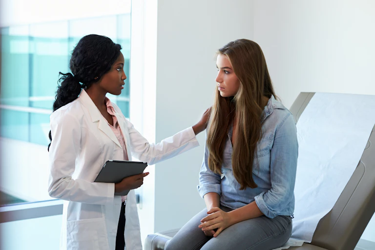 A doctor in a white coat speaks to a young female patient sitting on an examination table