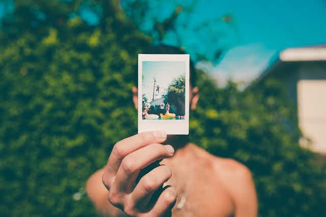 A photo of a shirtless man standing outside in the sunshine holding a faded Polaroid photo in front of his face