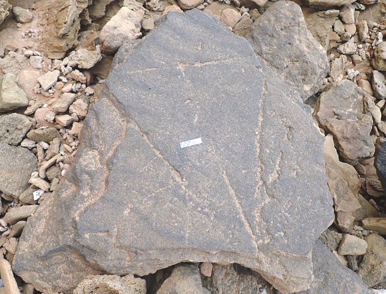 A large grey rock surrounded by smaller stones and coarse grains of sand. There are triangular carvings on the large rock.