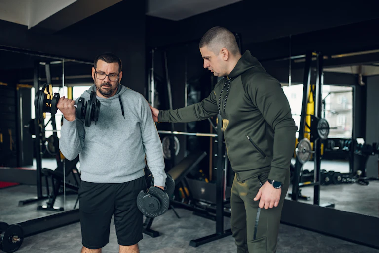 A middle aged man performs barbell curls in the gym while his trainer instructs him.