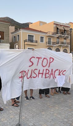 A group of people holding a large white sheet reading STOP PUSHBACKS in red. Only their feet are visible below the sheet.