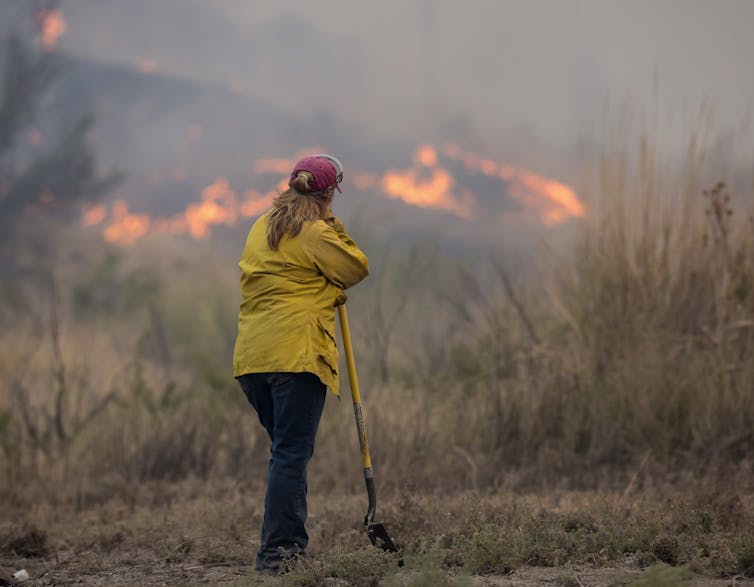 A woman in a yellow fireproof jacket leans on a shovel watching a wildfire on distant hills