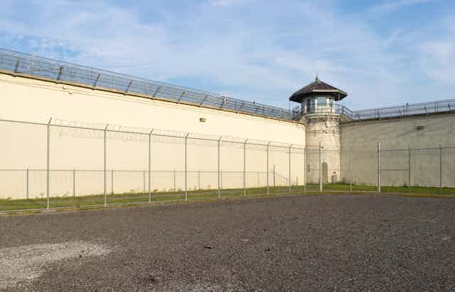 A prison yard is seen from the inside with a view toward two high walls that intersect at a watch tower.