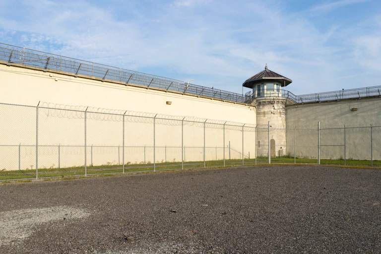 A prison yard is seen from the inside with a view toward two high walls that intersect at a watch tower.
