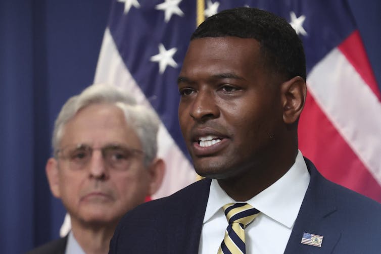 A Black man speaks in the foreground, with a white man and an American flag behind him