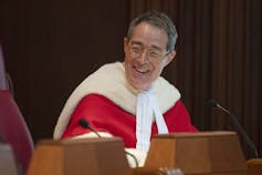 A man in a red robe with a white fur trim smiles sitting behind a desk.