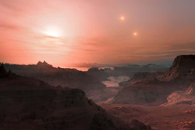 Artist impression of a mountainous landscape with a lake and possibly an ocean just before the horizon. One sun is just setting, but two other suns still appear in the sky. The sky is hazy and the sunset has turned it red.