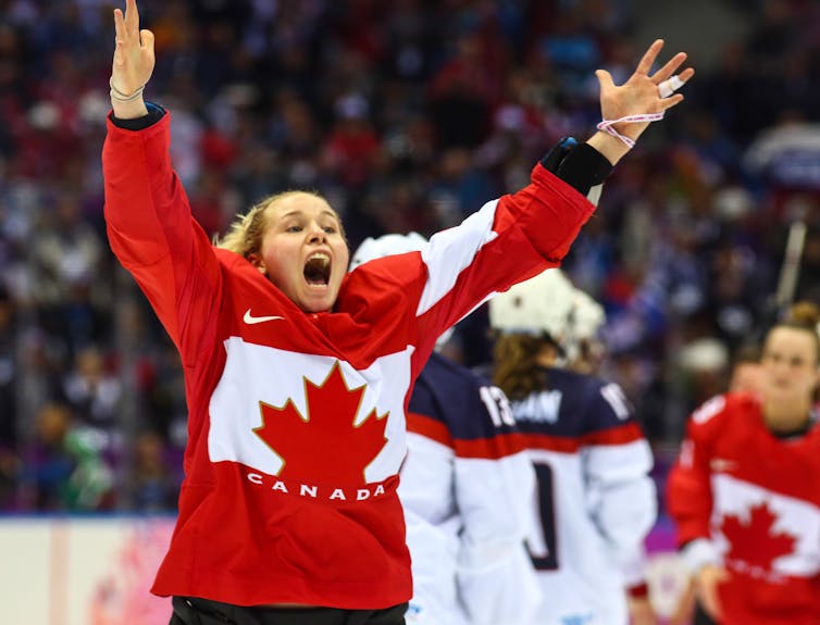 A woman wearing a red hockey jersey with the Canadian maple leaf raises her hands in celebration.