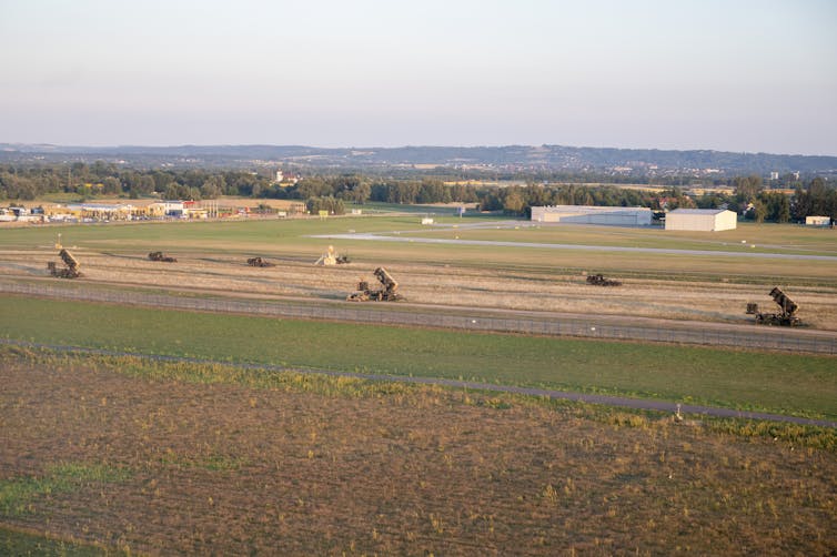 an aerial view of an airfield with an array of box-like structures in the foreground and hangers in the background