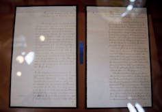 A hand-written document is seen behind glass.