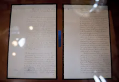 A hand-written document is seen behind glass.