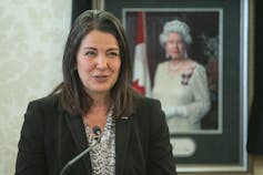 A dark-haired woman talks into a microphone, a framed photo of Queen Elizabeth behind her.