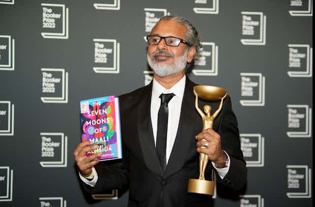 Man in suit with glasses holds book in one hand and a trophy in the other, against a Booker Prize 2022 backdrop