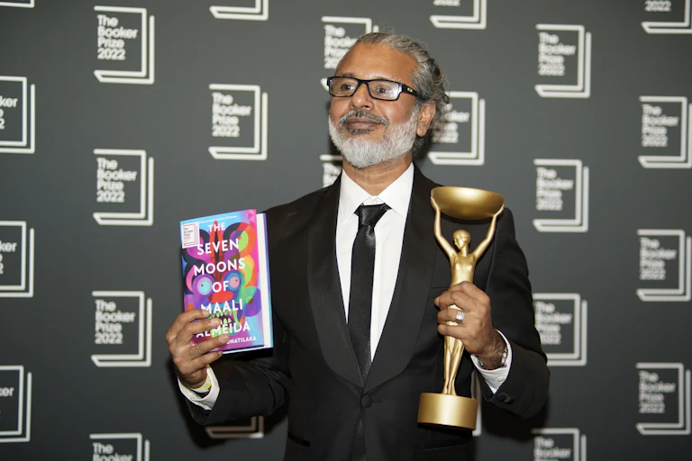 Man in suit with glasses holds book in one hand and a trophy in the other, against a Booker Prize 2022 backdrop