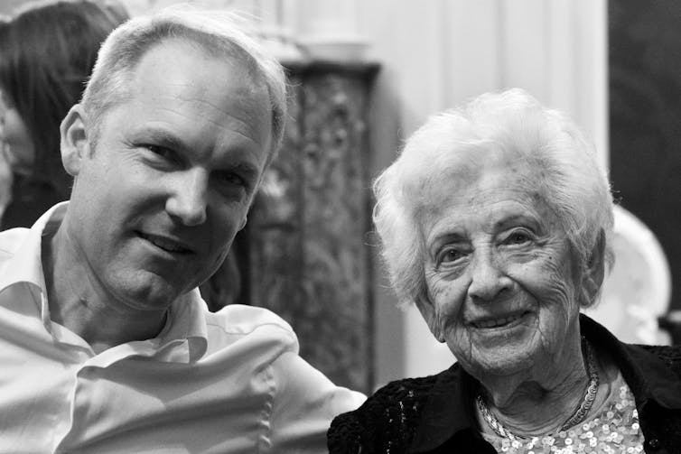 A man and an older woman, smiling at the camera. He is in a white shirt and she wears a sequinned top under a cardigan