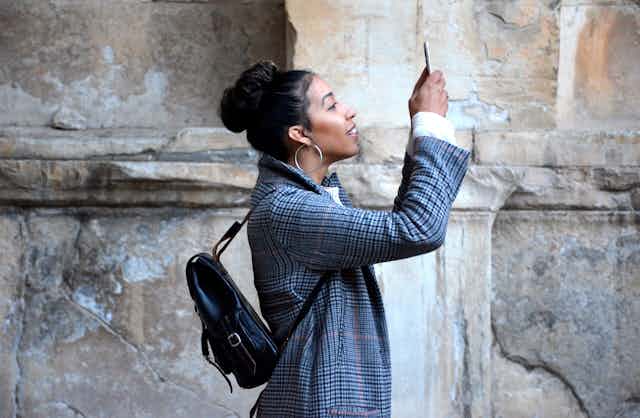 A woman with dark hair in a blazer taking a selfie in front of a sandstone wall