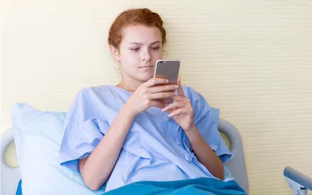 A young woman in a hospital gown looking at her smartphone in a hospital bed