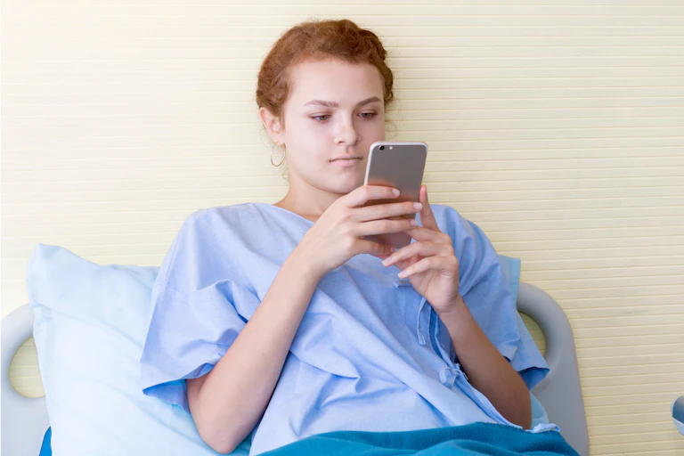 A young woman in a hospital gown looking at her smartphone in a hospital bed