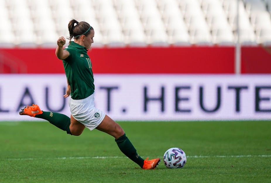 Katie McCabe, wearing a green Ireland jersey, white shorts and neon orange shoes, approaches a stationary football to take a free kick