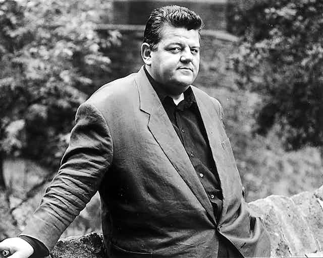 A black and white image of Robbie Coltrane standing by a wall.