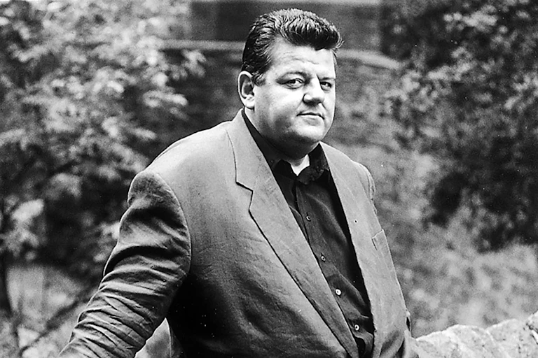 A black and white image of Robbie Coltrane standing by a wall.