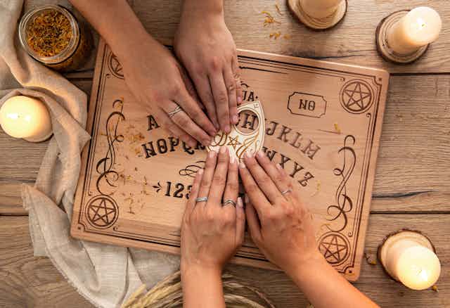 Photo from above of two people's hands using a Ouija board, surrounded by candles