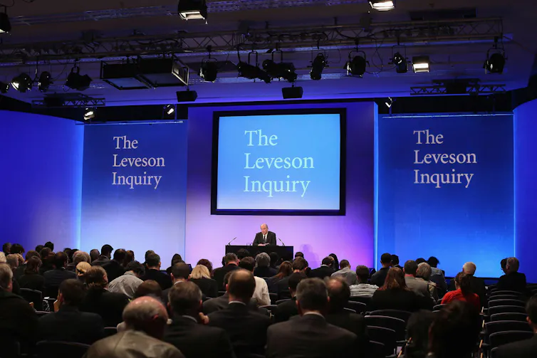 Man sits at table in front of seated crowd under sign reading 'The Leveson Inquiry'.