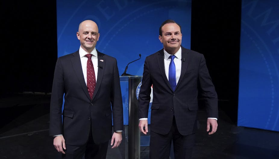 Two white men in suits -- one with a red tie, the other with blue -- stand in front of a lectern on a stage.