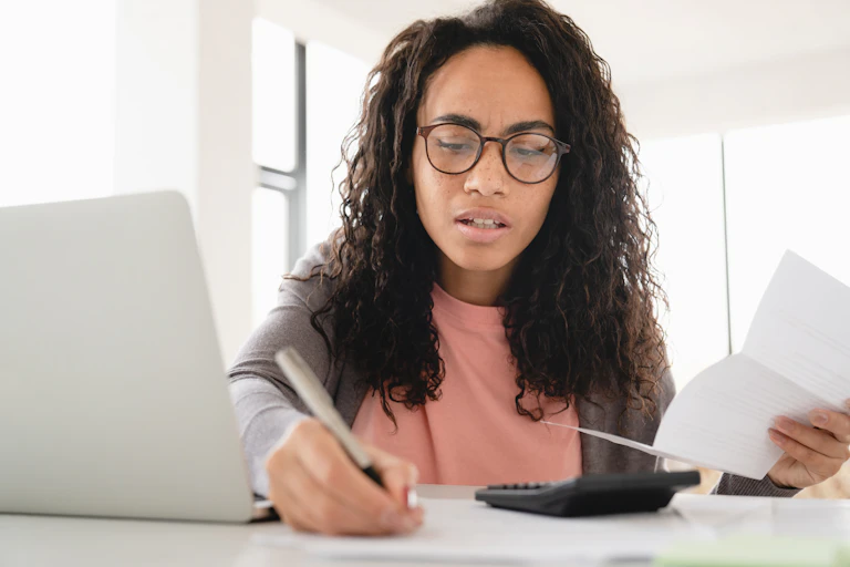 A woman checking her finances with a pen, laptop, bill and calculator.